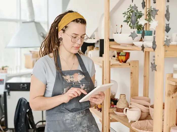 Woman artist checking orders on a tablet in her pottery studio, creating handmade products for towndirectoryworks.com